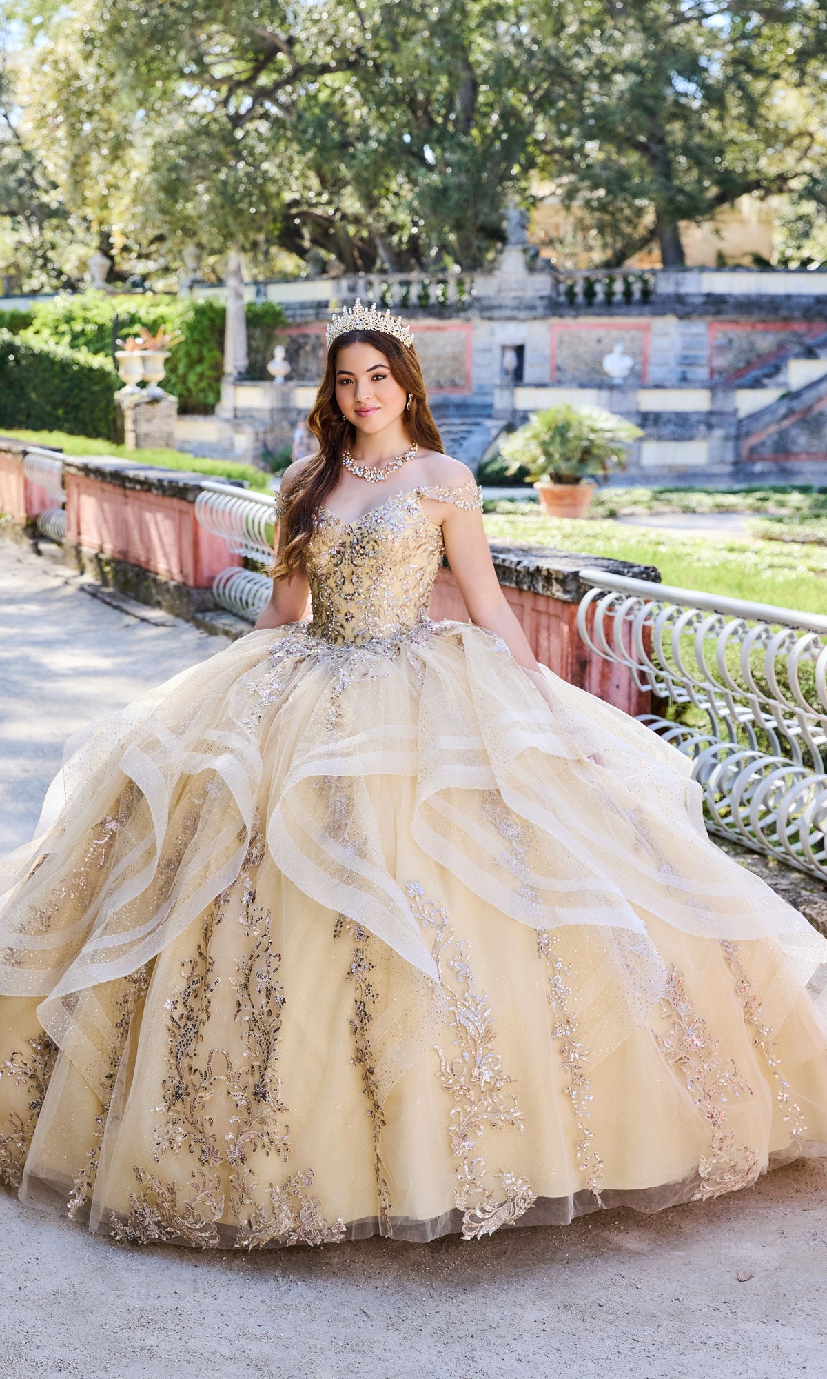 A young woman wearing the Quinceanera Dress PR30117 By Princesa and a tiara stands outdoors on a garden path, surrounded by greenery and stone structures.